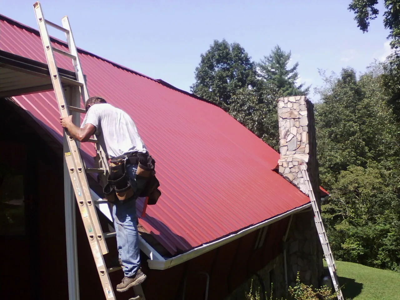 Roofer on a ladder beside a red metal roof and stone chimney — Soffit & Fascia Repair by Roofing Brothers in Blytheville